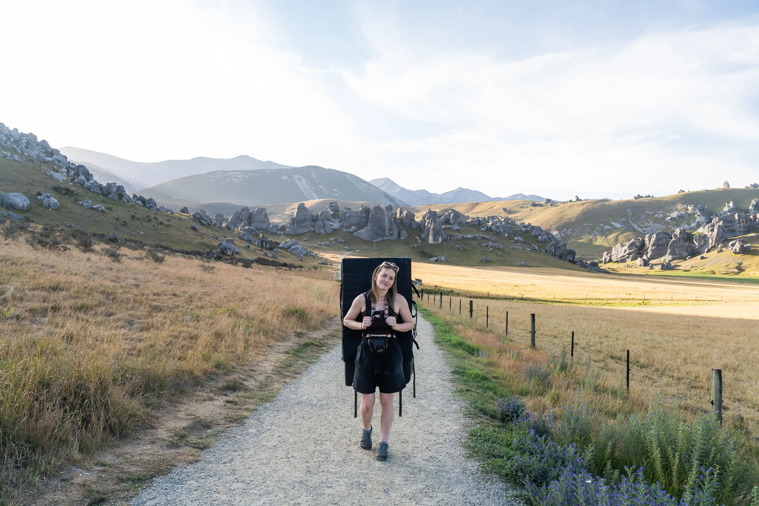 A woman hiking on a gravel trail in a scenic grassy mountain landscape, carrying a large backpack and holding a camera, with rocks and rolling hills in the background.