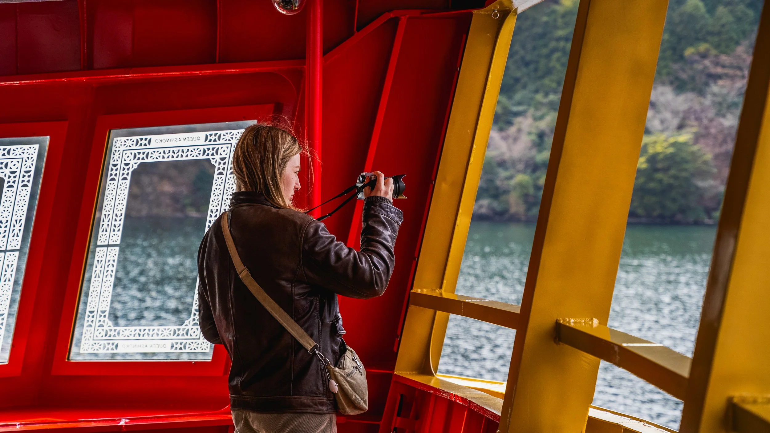 A woman taking a photo with a camera on a boat, with water and a forested shoreline in the background.