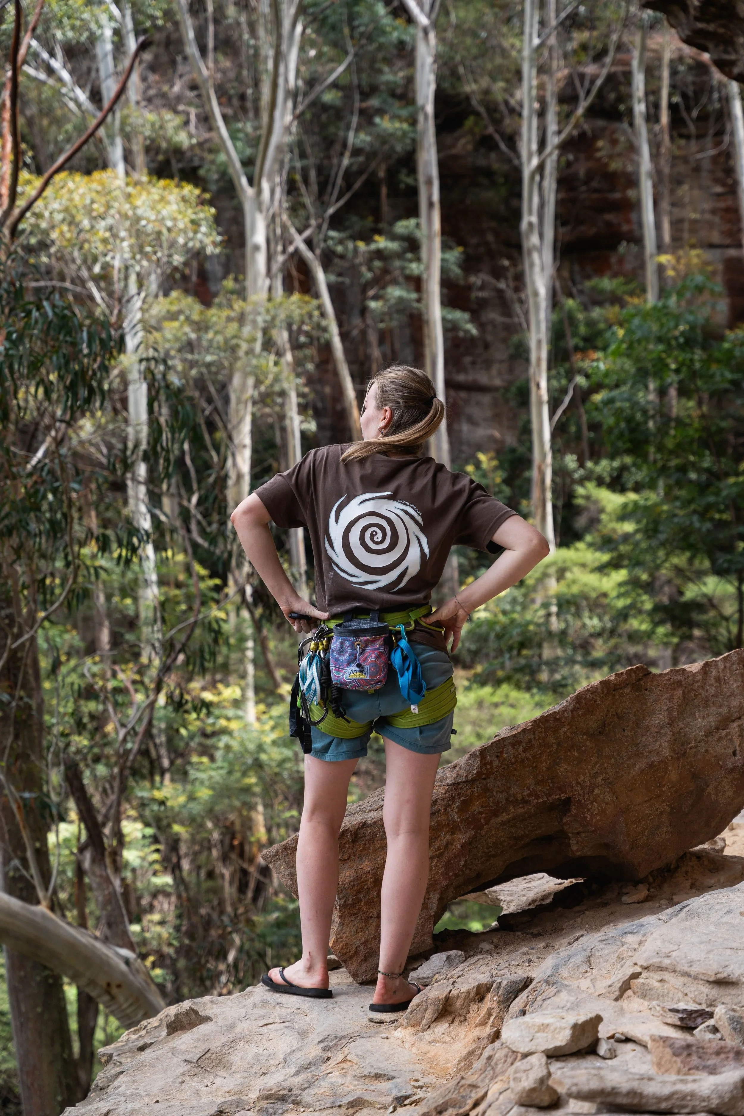 A woman wearing a brown t-shirt, shorts, and flip-flops stands on a rock in a forest, looking at the trees and rocky terrain ahead.
