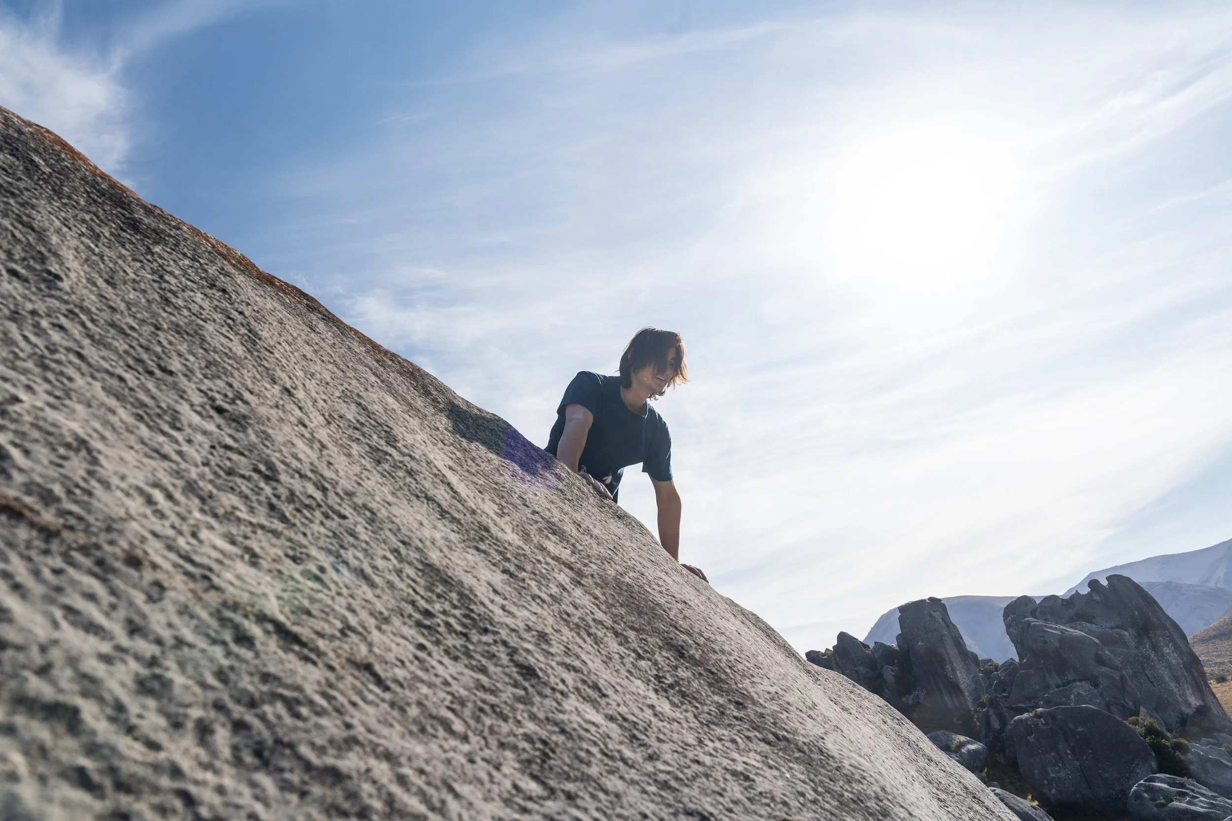 A person climbing incline on a rugged outdoor rock surface with mountains and a bright sky in the background.