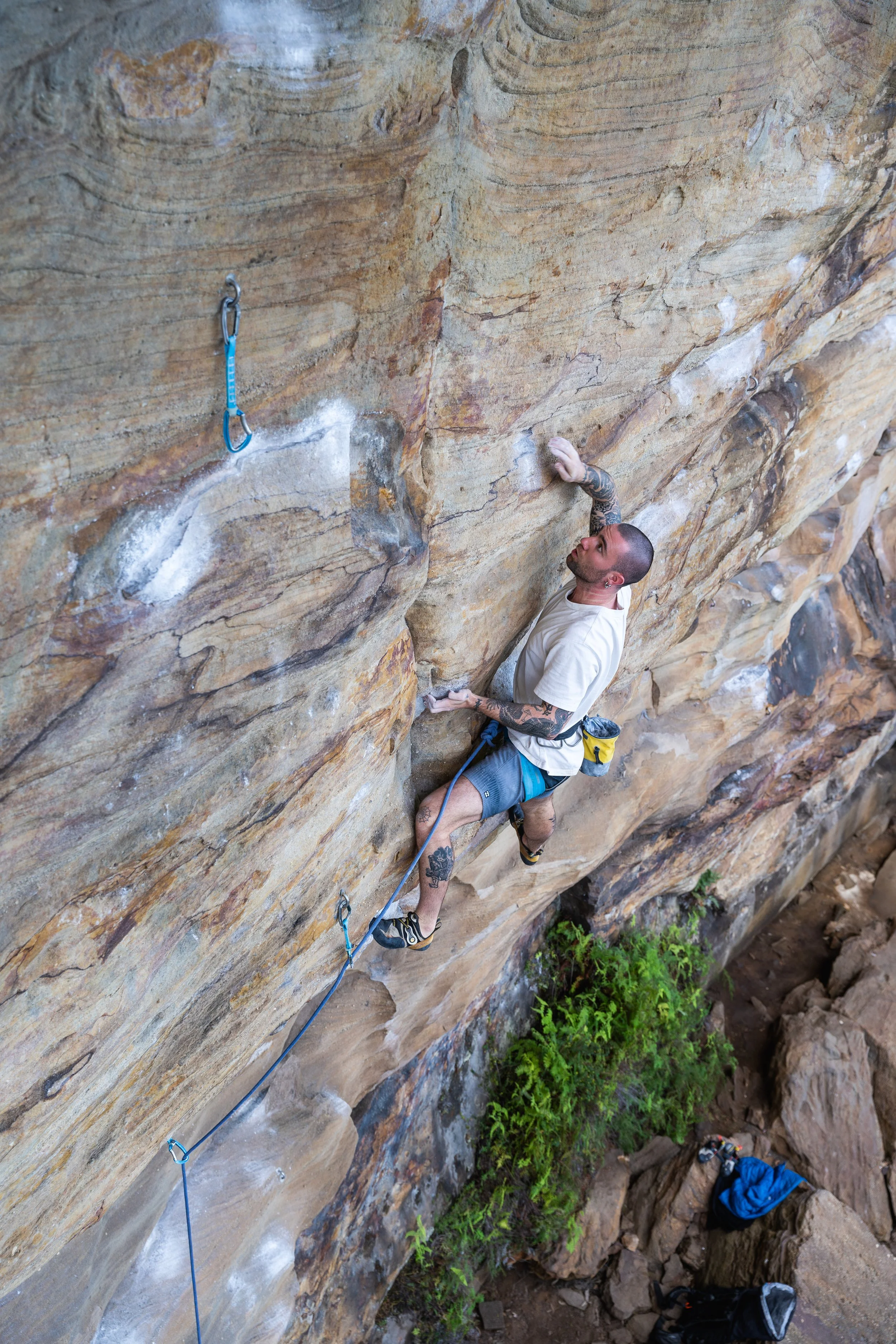 A man is climbing a steep, rocky outdoor cliff face with climbing gear, including a harness, quickdraws, and a climbing rope, with another person below at the base of the cliff.