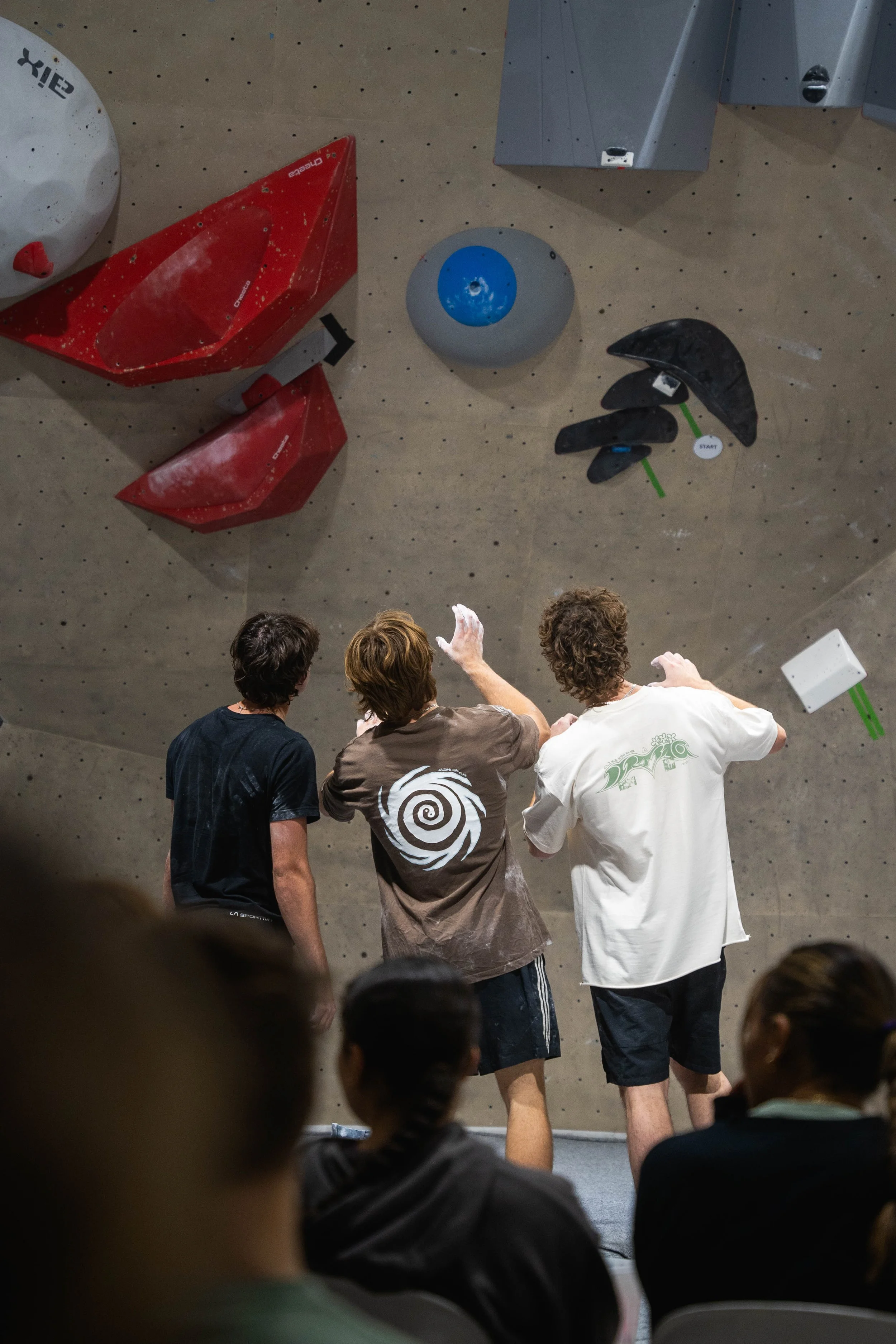 Three young men stand in front of an indoor rock climbing wall, preparing to climb or demonstrating techniques, with climbing holds of various shapes and colors on the wall and an audience seated in front of them.