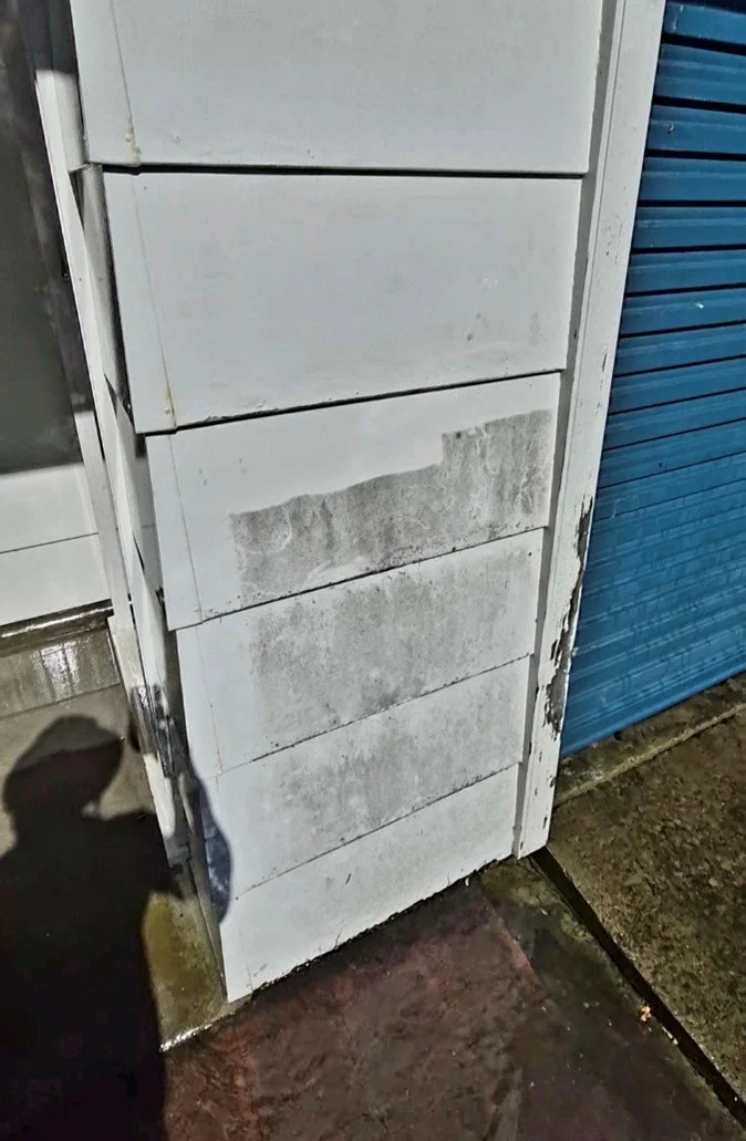 A white panel on a building's exterior shows signs of dirt and mold, with a blue rolling shutter door next to it.