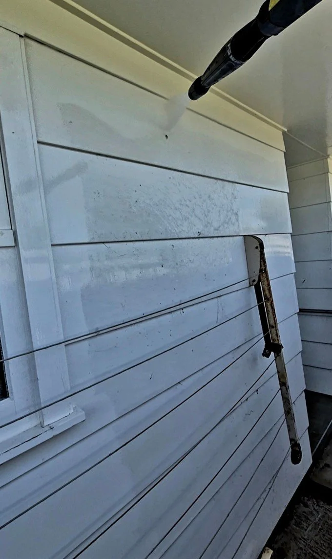 A water hose is spraying water onto the siding of a house. A long metal pry bar leans against the house below the hose. The house has white horizontal vinyl siding and a small window is visible on the left.
