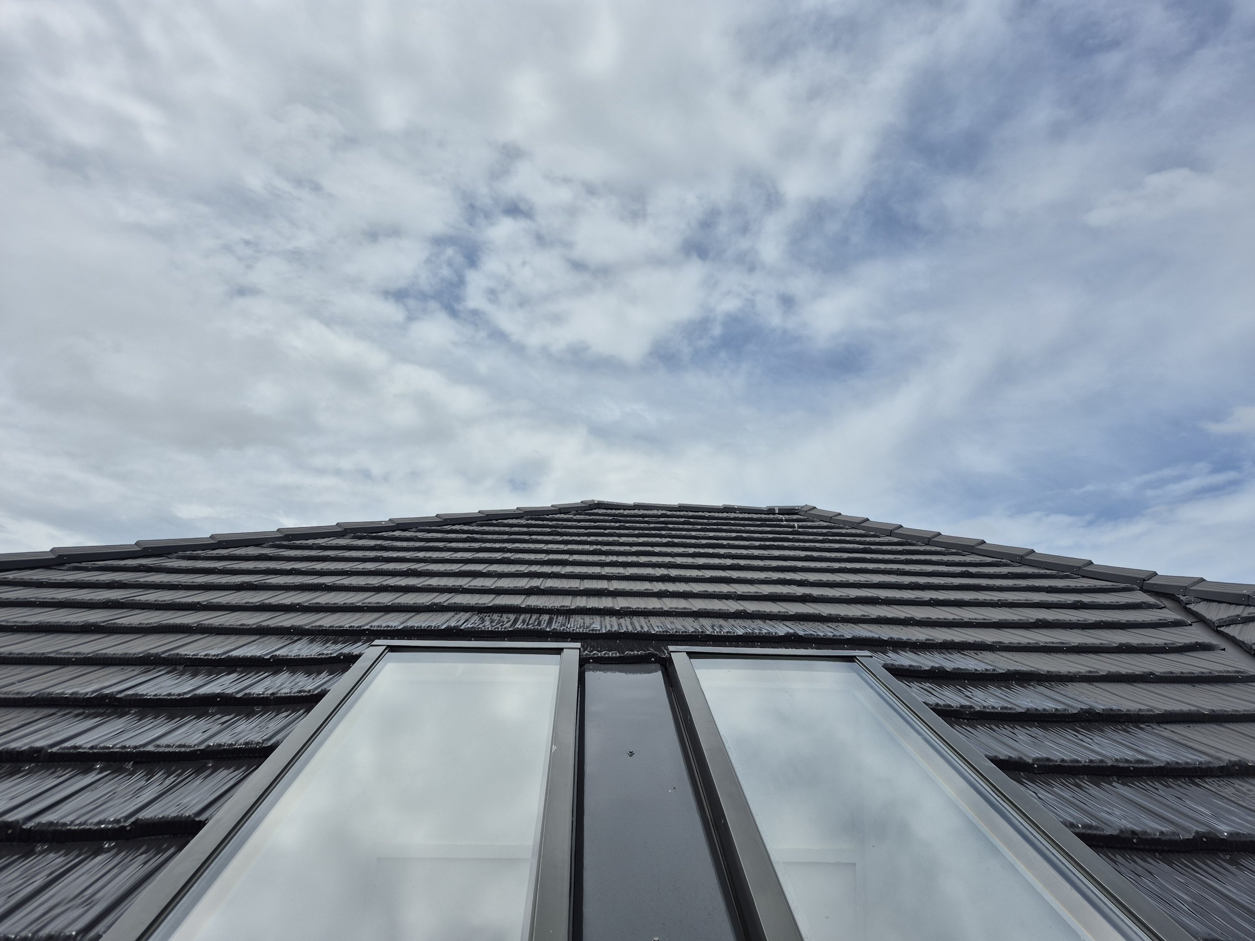 Low-angle view of a house roof with dark shingles and a glass window, under a partly cloudy sky.