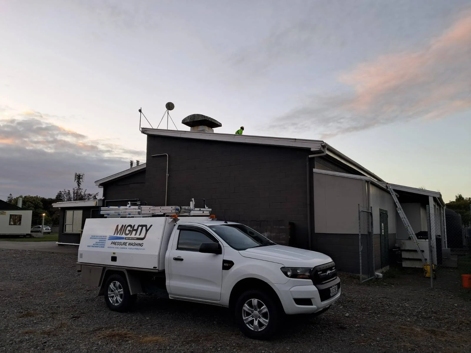 A white service truck labeled 'MIGHTY Pressure Washing' parked in front of a dark-colored building with a ladder leading to the roof, where a worker in a green shirt is working at sunset.