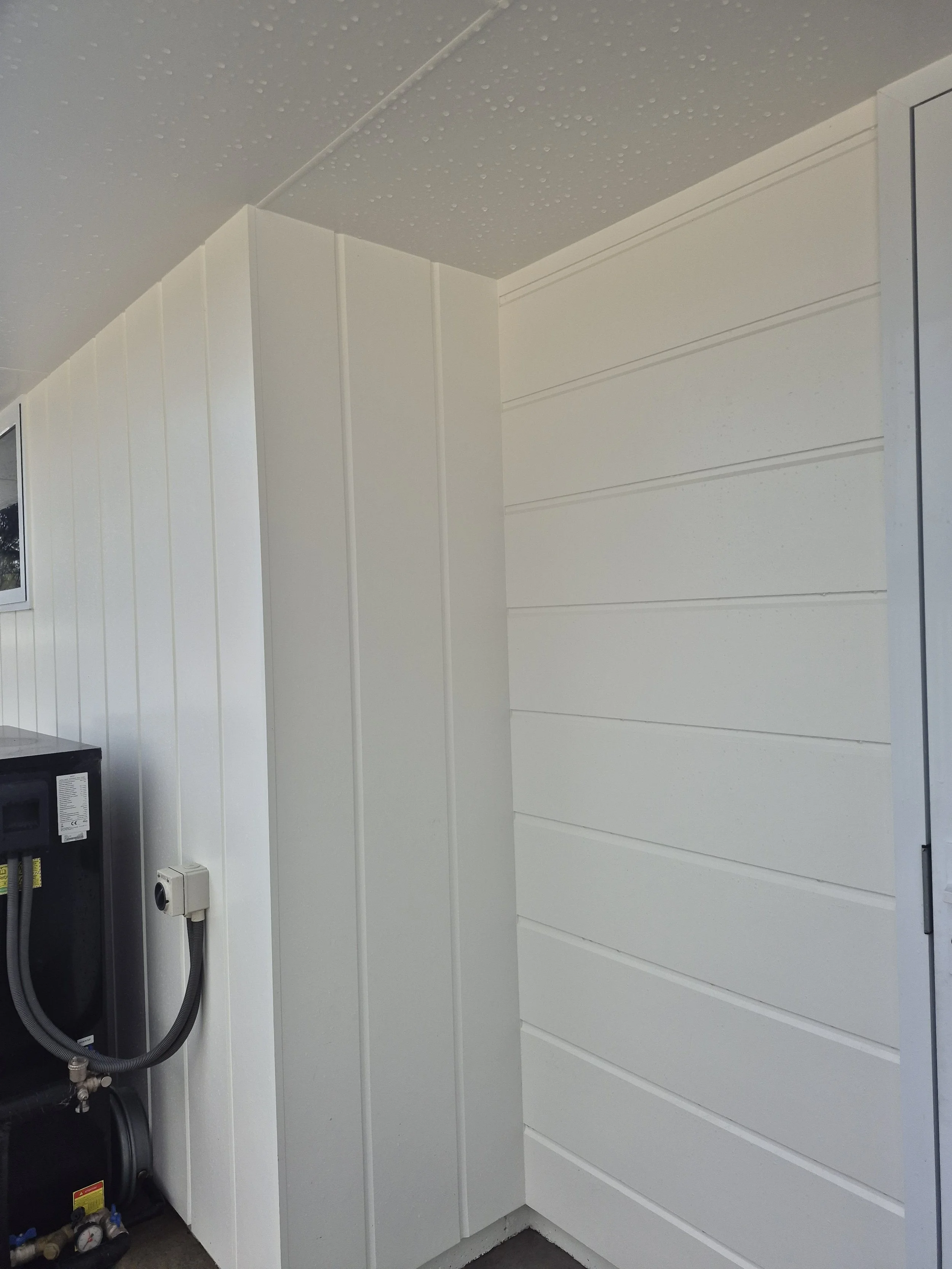 A corner of an indoor room with white vertical paneling on the wall, a small section of black equipment on the left, and part of a door or cabinet on the right. There is a small rectangular object attached to the wall near the equipment.