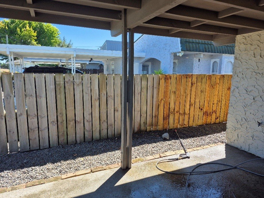 View of a backyard with a partially painted wooden fence, a gravel ground, and a house in the background, taken from a covered patio area with a concrete floor and a garden hose.