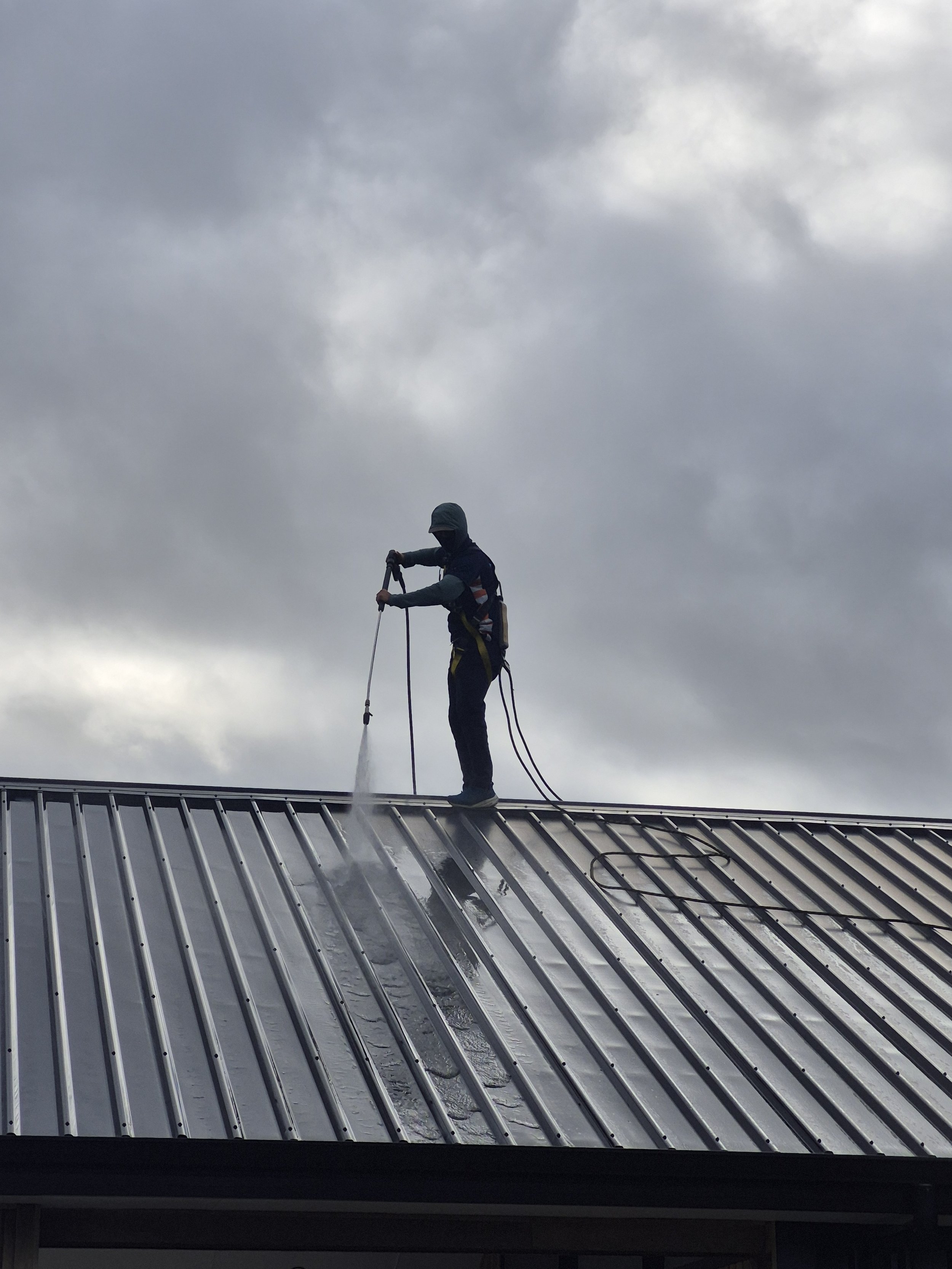 A person wearing safety gear cleaning a metal roof with a high-pressure washer against a cloudy sky.
