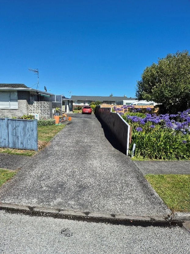 A residential driveway leading to a house with a red car parked near the home, surrounded by a garden with purple flowers and a clear blue sky.