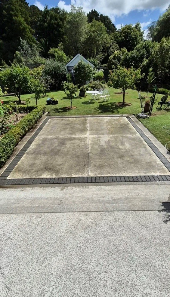 Empty concrete driveway with brick border leading to a lush green backyard with trees, a small shed, outdoor furniture, and garden tools.