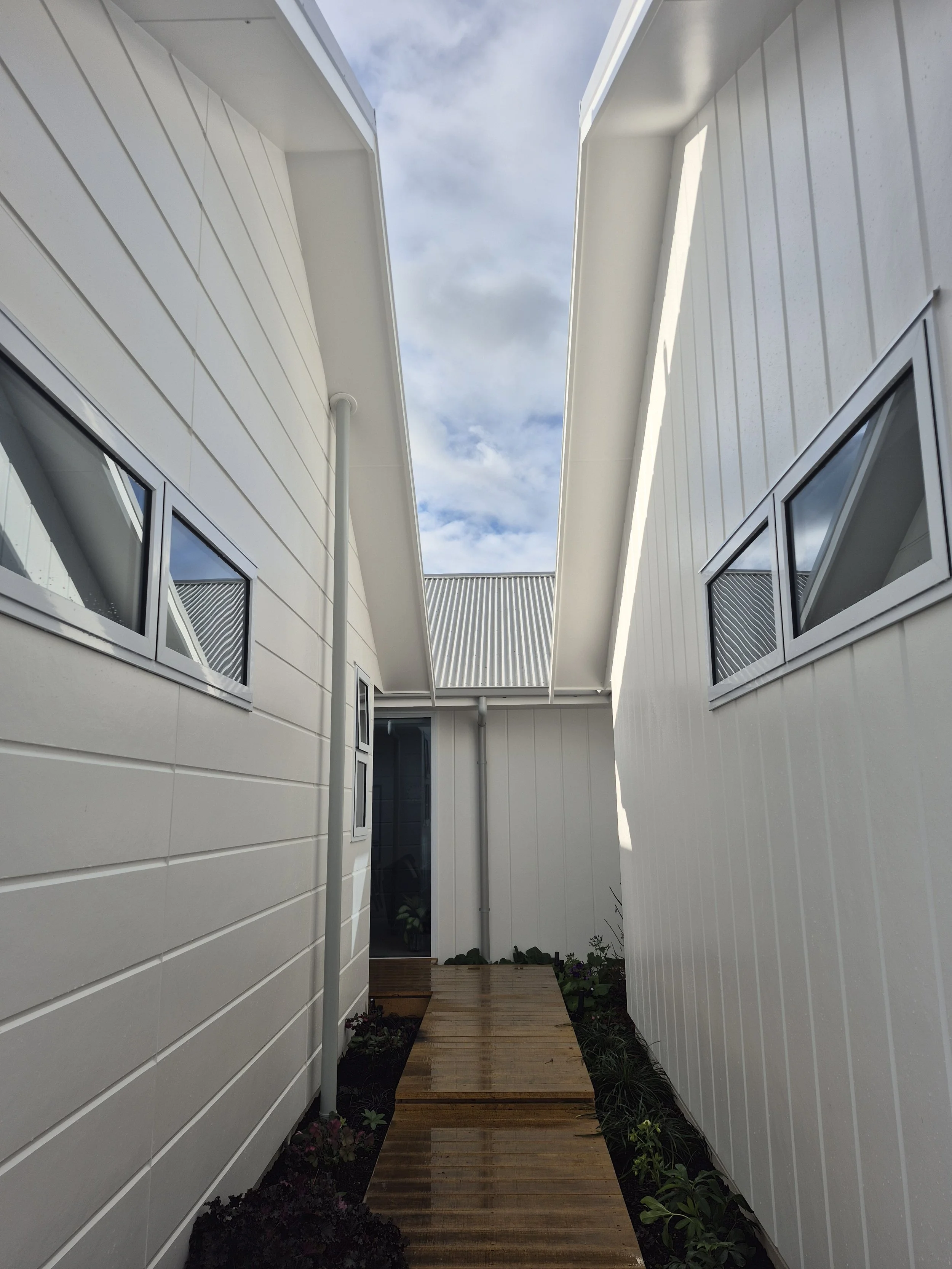 Narrow wooden pathway between two white modern houses with horizontal siding, small windows, and a cloudy sky overhead.