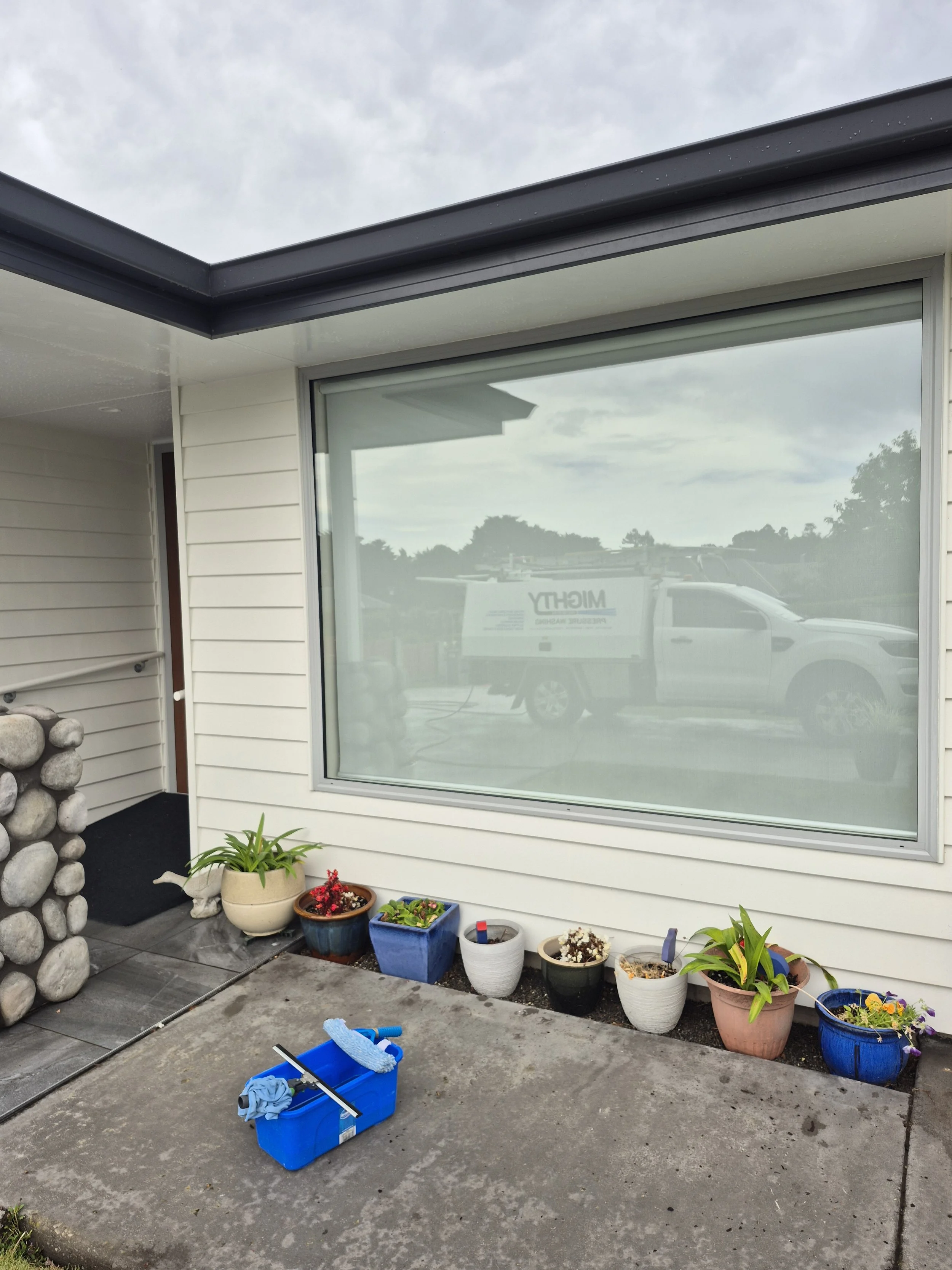 Front porch area with potted plants, a cleaning bucket, and a large window reflecting a parked vehicle and cloudy sky.