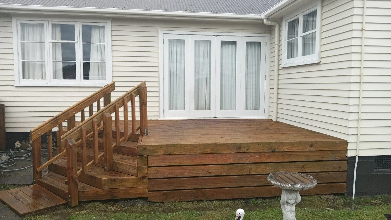 Wooden deck attached to a house with white siding, featuring a railing, steps, and a sliding glass door, with a small bird bath and a lawn in the foreground.
