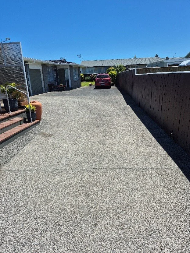 A gravel driveway leading to a garage with a red car parked near a house, with a dark fence on the right side and house with plant pots on the left, under a clear blue sky.