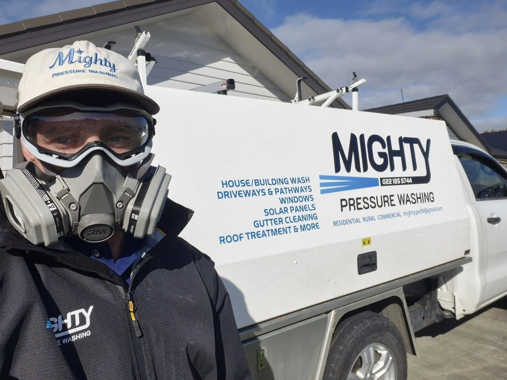 A man wearing a cap and respirator mask taking a selfie in front of a white work truck with the company name 'Mighty' and services listed on the side, including house and building wash, driveway and pathway cleaning, window cleaning, solar panel clea