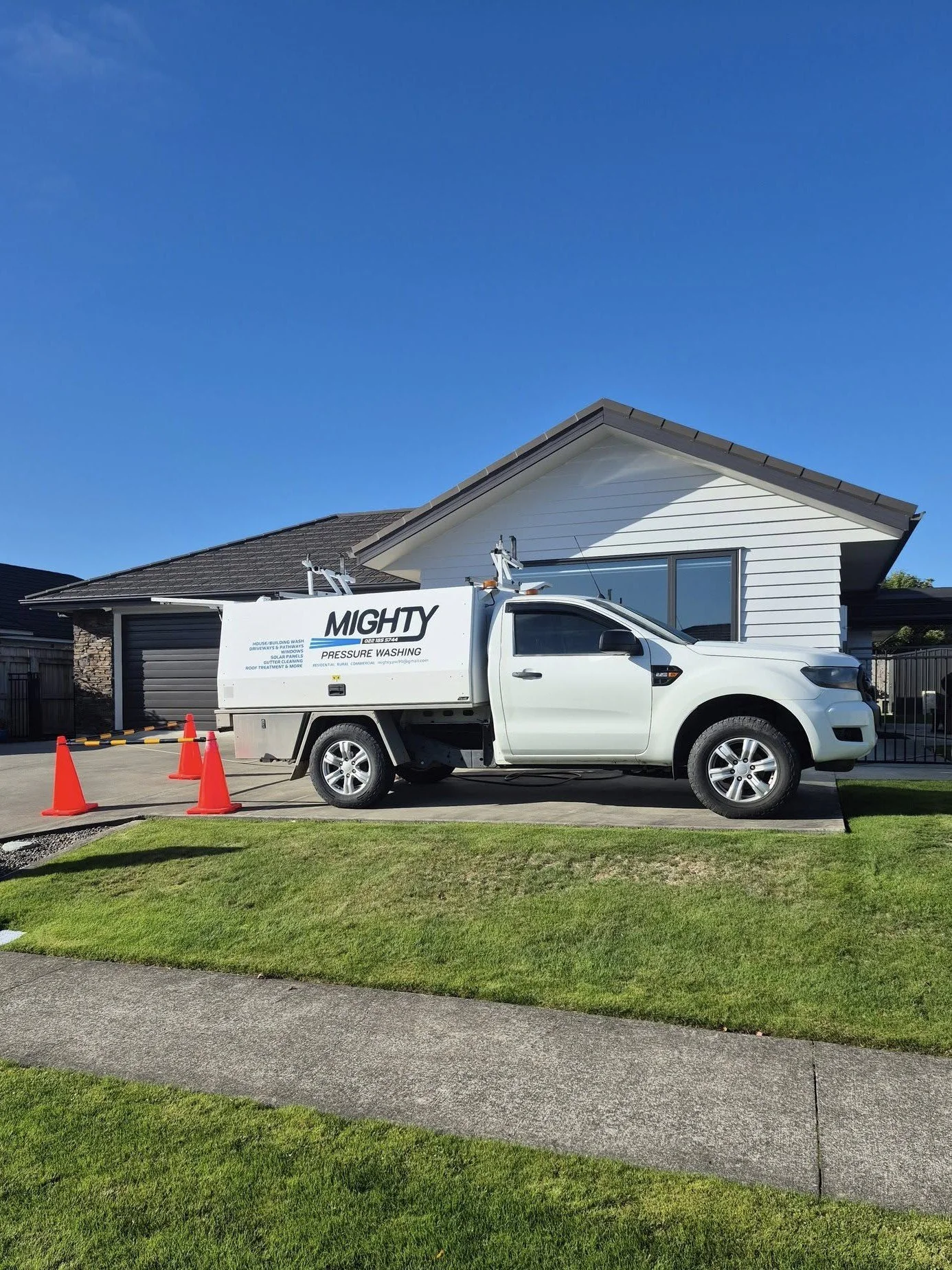 White truck with 'MIGHTY PRESSURE WASHING' logo parked in driveway of a house with white siding and a gray roof, surrounded by orange cones and safety barriers on a sunny day.