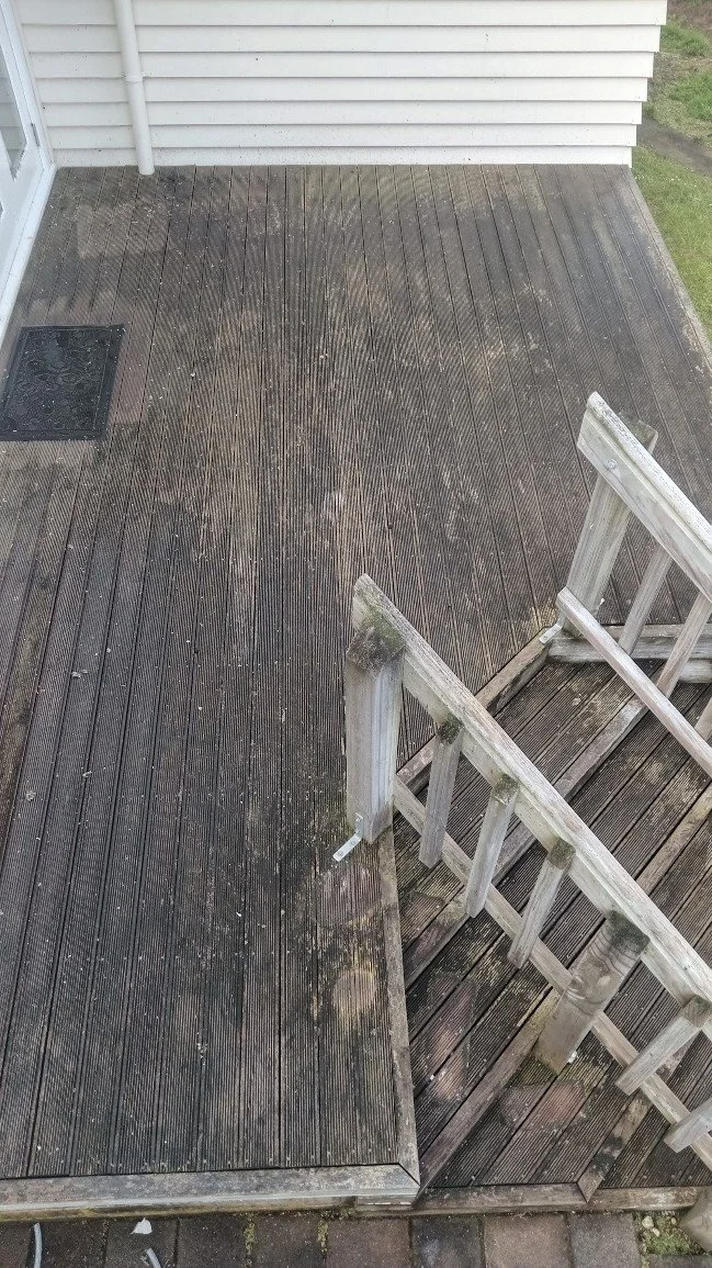 A wooden deck with some dirt and moss, attached to a house with white vertical siding. A staircase leads down from the deck. There is also a small black doormat near a door.