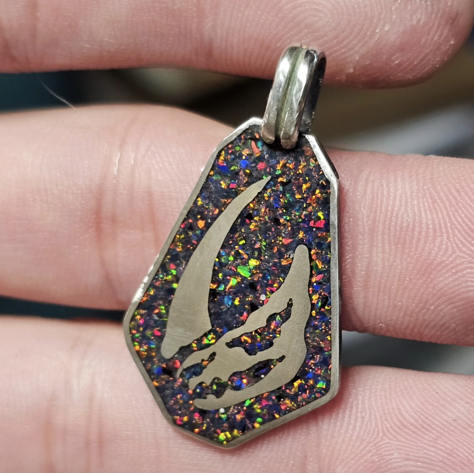 A close-up of a pendant with a silver border, containing a colorful, glittery background and a silver-colored silhouette of a hand making the shaka sign.