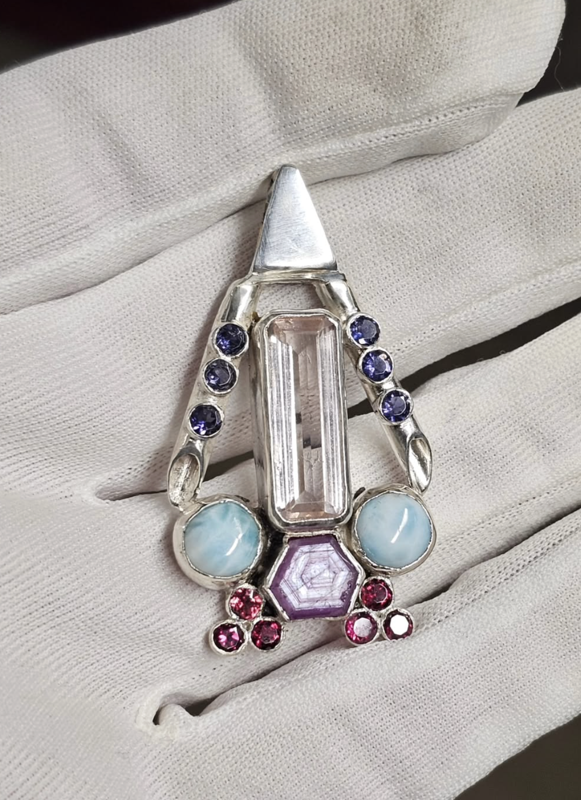 A silver ring with colorful gemstones including a large rectangular pink gemstone, two blue cabochon stones, a purple hexagonal stone, and numerous small pink and purple stones, held on a white cloth background.