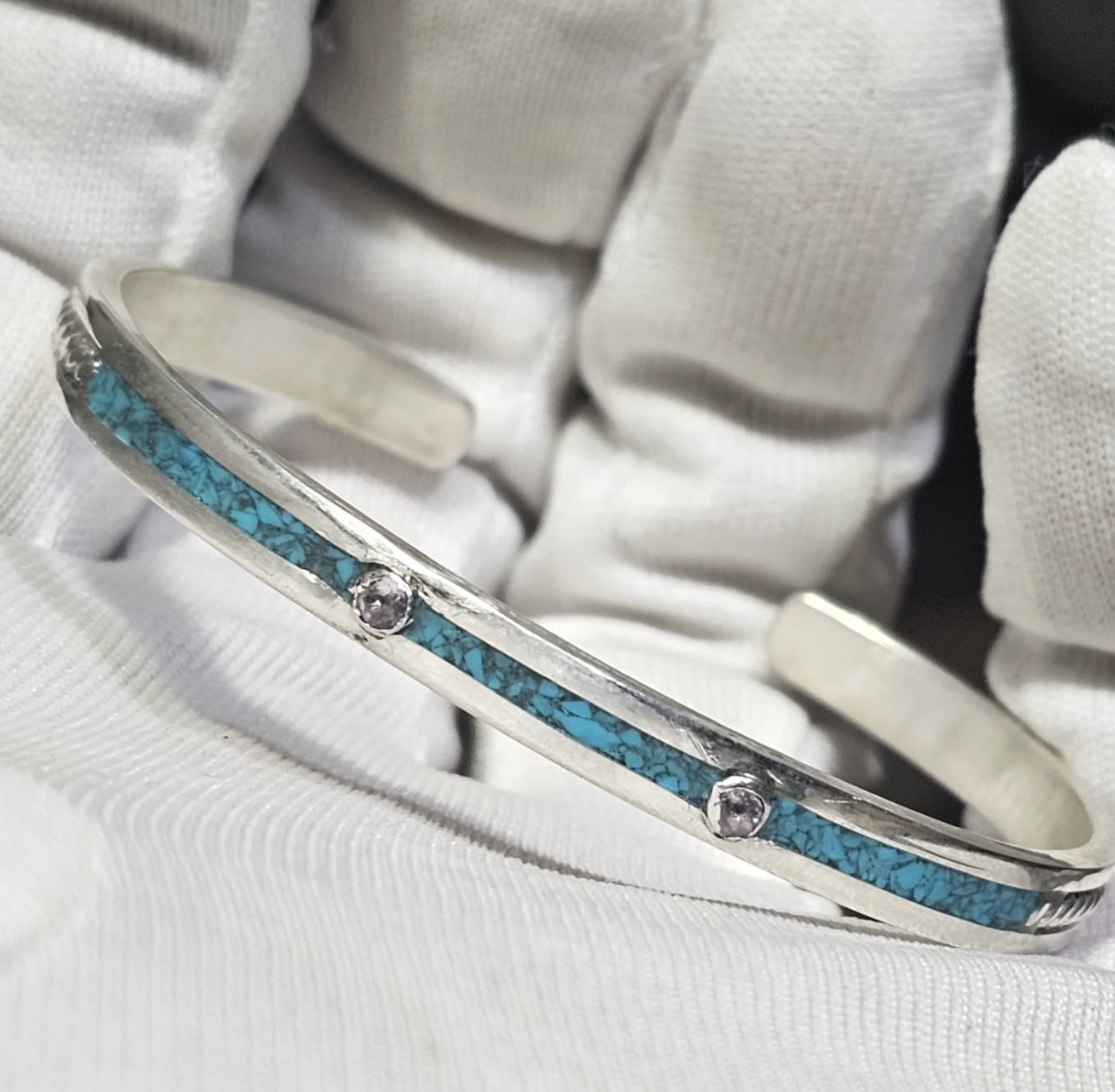 A silver bracelet with turquoise inlay and small round gemstone accents, held by white fabric gloves.