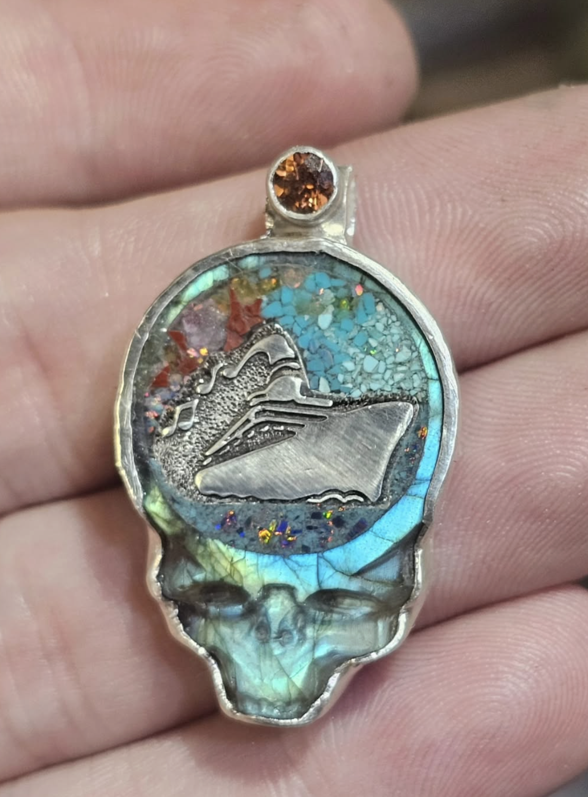 A necklace featuring a round pendant with a colorful, glittery background, a silver carving of a mountain, and an iridescent gemstone at the top.