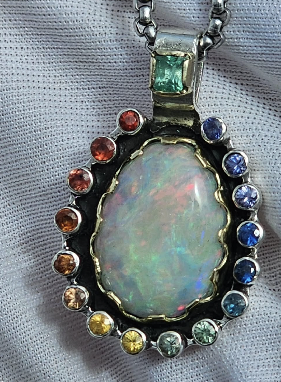 Close-up of a pendant with a large oval opal gemstone surrounded by multicolored smaller gemstones, attached to a silver chain with a rectangular emerald gemstone at the top. The background is a textured white fabric.