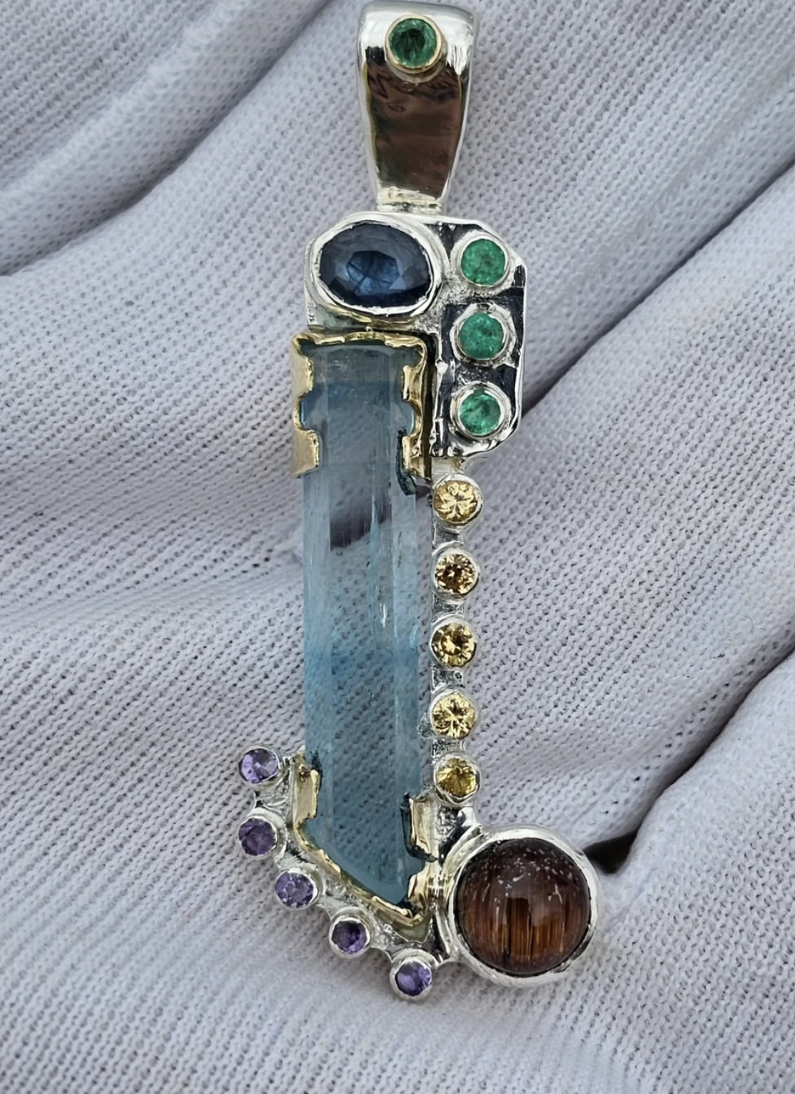 A multicolored gemstone pendant having various shapes and sizes with a large blue rectangular gemstone in the center, surrounded by small round gemstones in green, yellow, purple, and a large brown round gemstone at the bottom, all set in a silver an