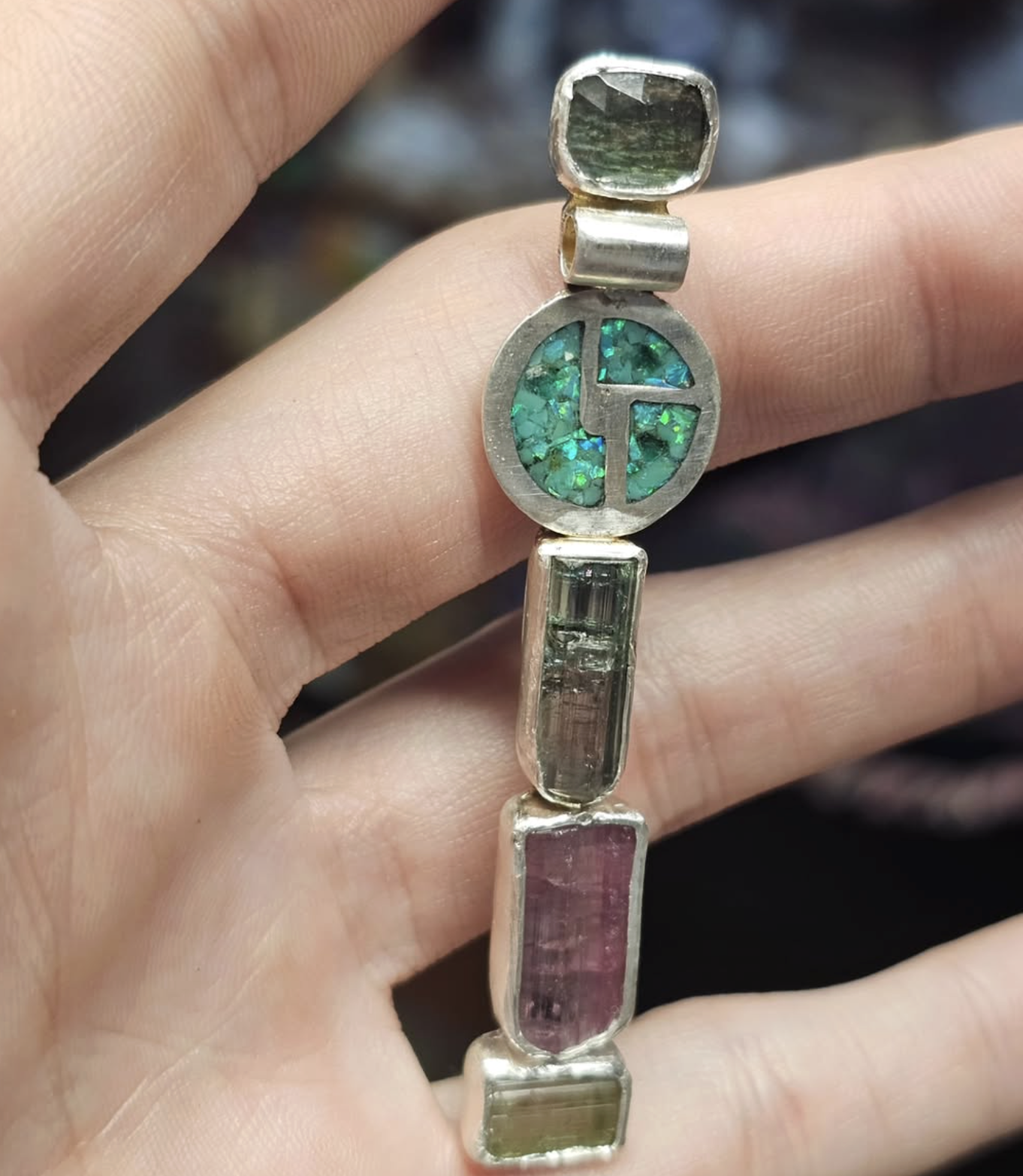 Hand holding a silver bracelet with multicolored gemstones, including green, pink, purple, and clear stones.