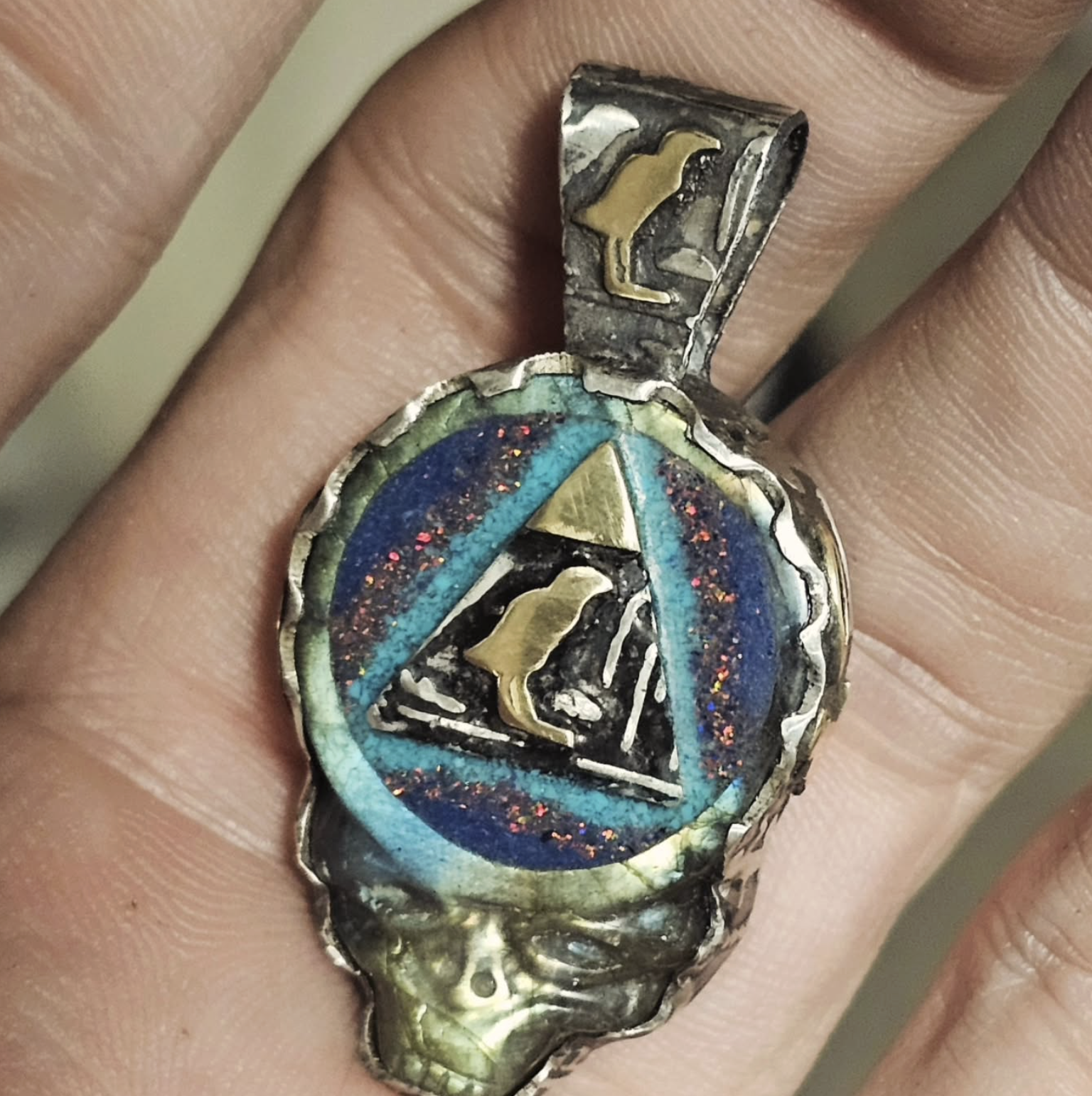 A close-up of a ring with a colorful, enamel inlay featuring a triangular symbol and a bird silhouette inside a circle, adorned with metallic skull details on the band.