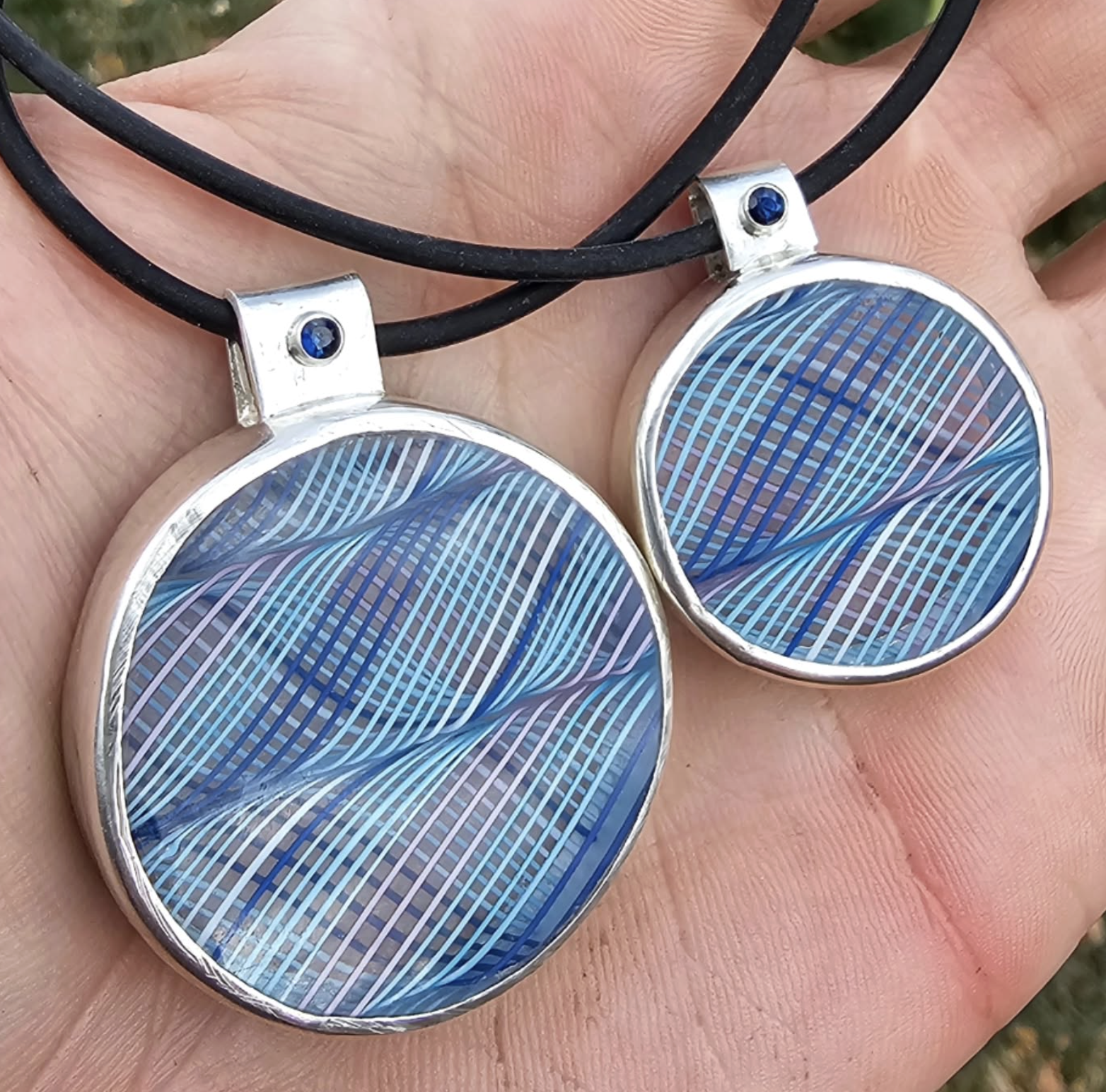 Matching jewelry set with round pendants featuring blue and purple abstract line patterns, attached to black cords with small blue stones at the top.