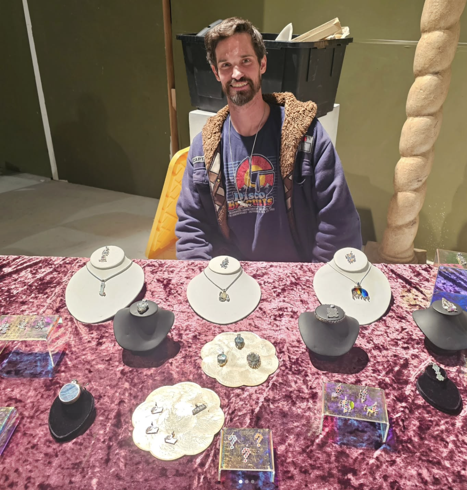 Man sitting behind a jewelry display table with necklaces and rings, covered with a pink velvet cloth, at an indoor event.