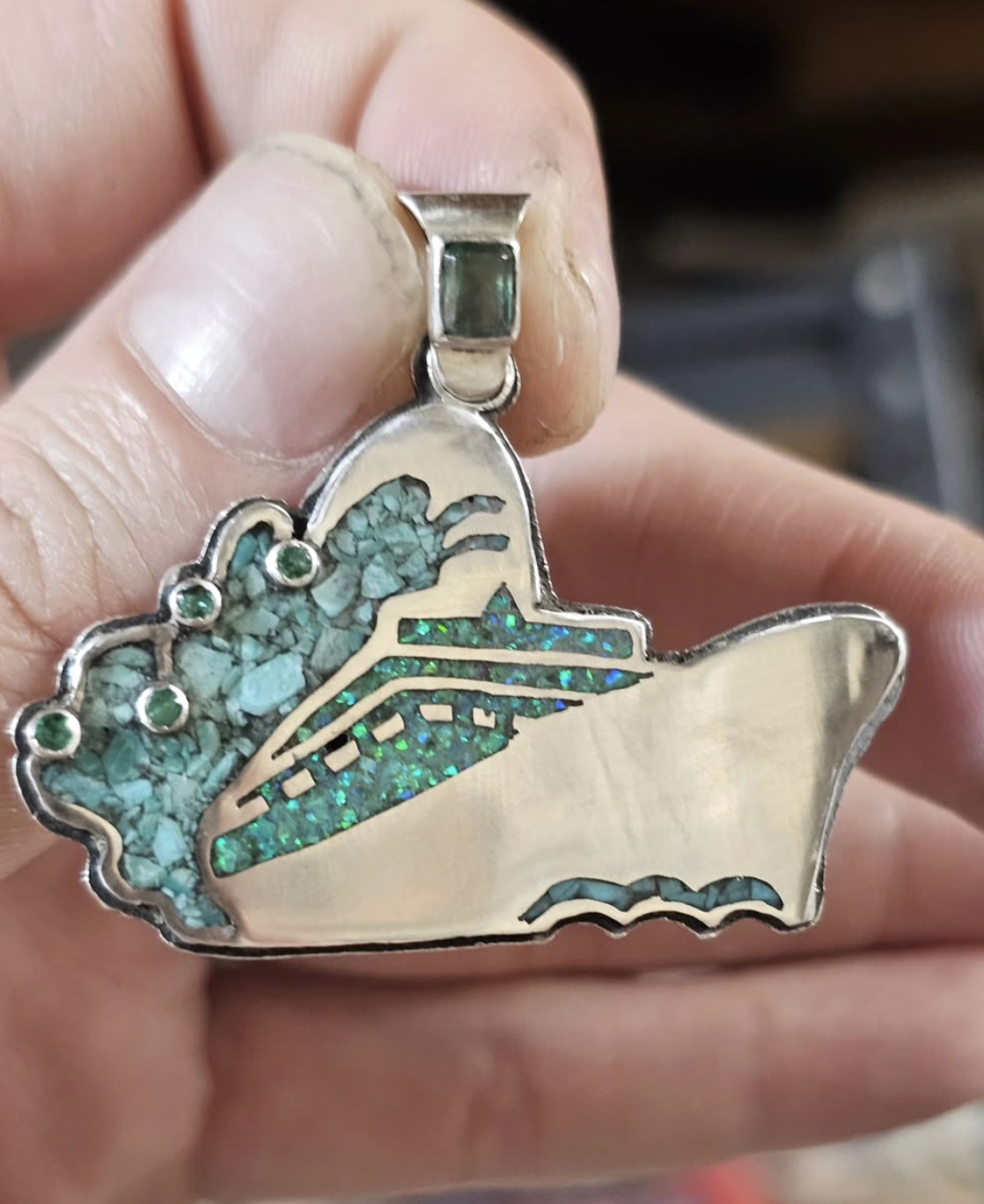 A silver church bell pendant attached to a hand, featuring a decorative butterfly design with turquoise and iridescent inlays.