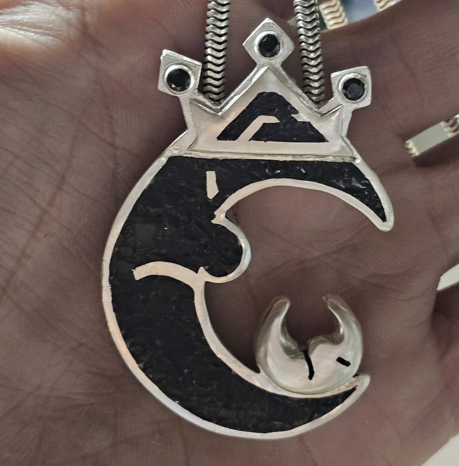 A silver necklace with a pendant featuring a crescent moon and face design, topped with a crown-like structure with three small black stones and a black mountain symbol, hanging from a silver chain.