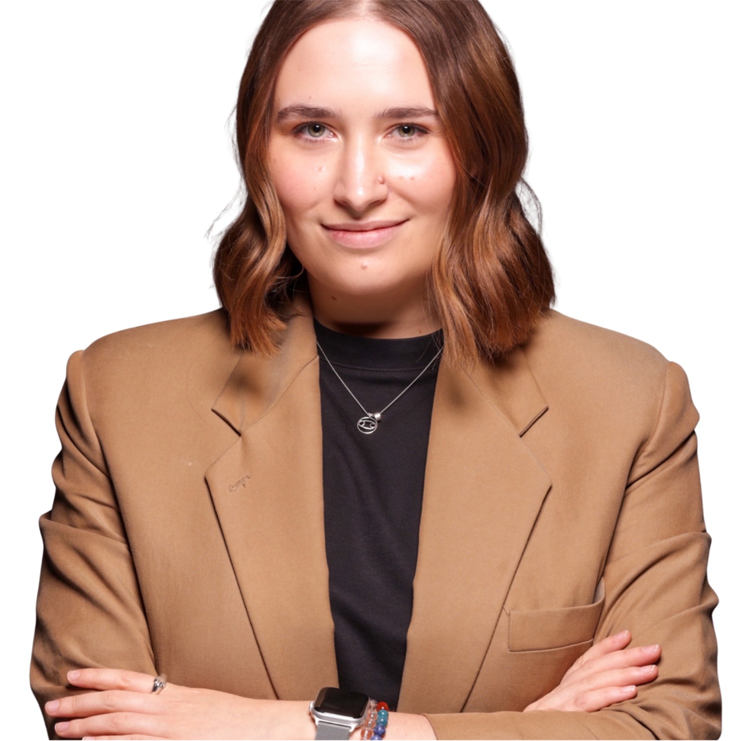 A woman with shoulder-length light brown hair and light skin, wearing a tan blazer over a black top, sitting with her arms crossed, smiling softly, wearing a necklace and a smartwatch with bracelets on her left wrist.