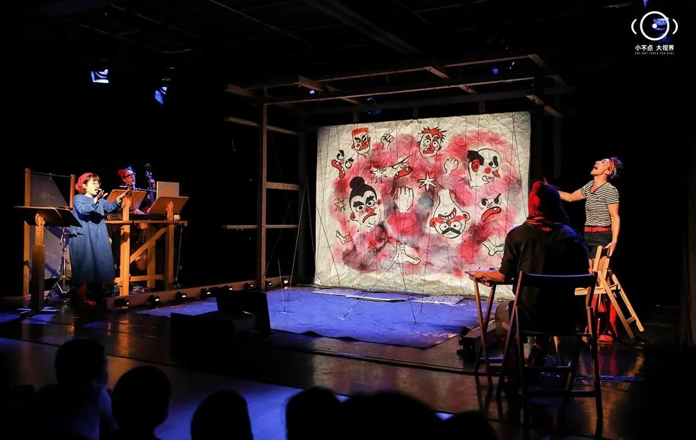 A theatrical performance on stage with puppetry and actors. The stage features a painted backdrop with various expressive faces. On the left, two children are operating puppets or marionettes, while on the right, two actors are engaged in a scene wit