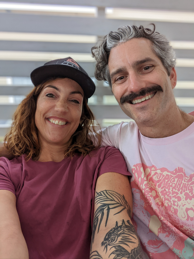 A man and woman smiling for a selfie, with the man on the right and the woman on the left. The woman has wavy brown hair, is wearing a black cap, and a maroon T-shirt. The man has curly gray hair, a mustache, and is wearing a light pink T-shirt with 