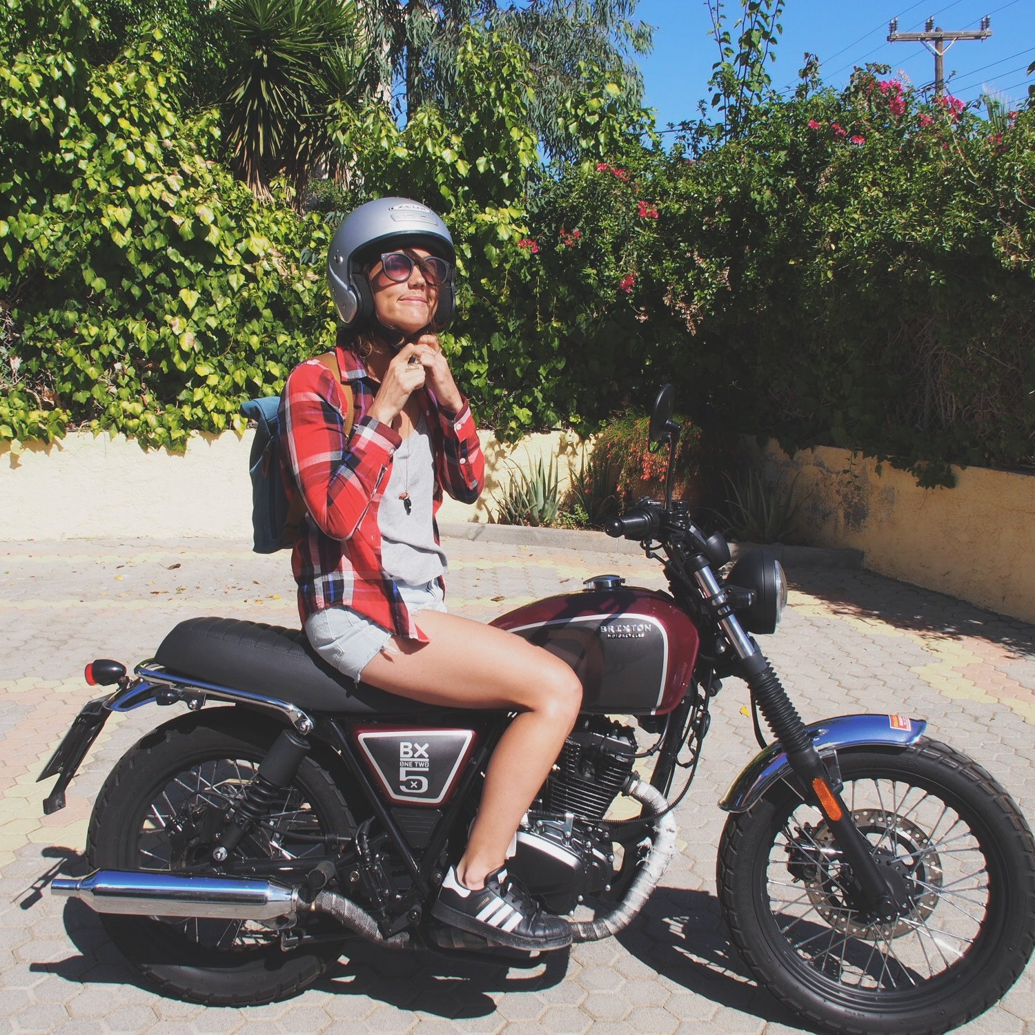 A woman sitting on a motorcycle wearing a helmet, sunglasses, a red plaid shirt, gray shorts, and sneakers, with greenery in the background.