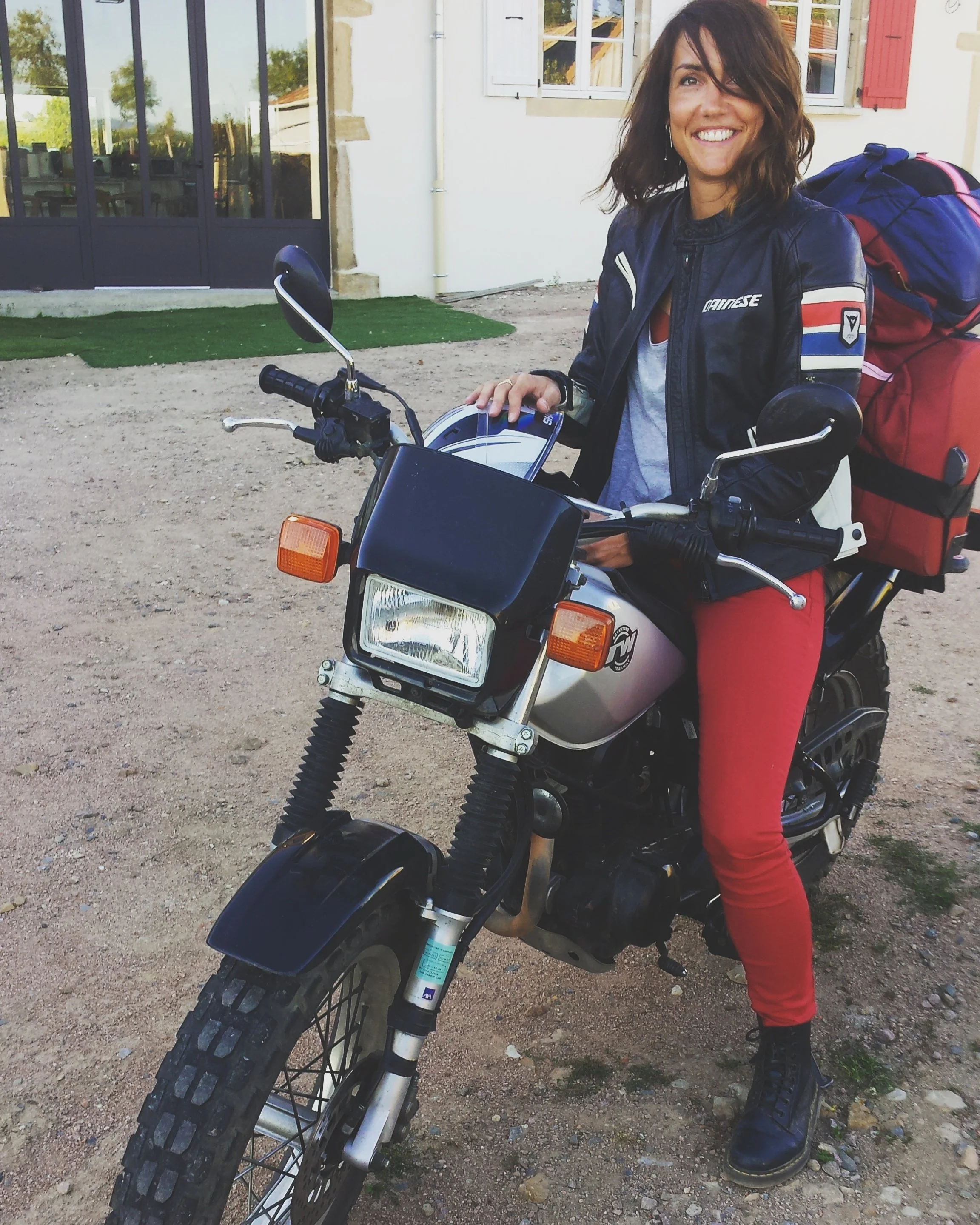 A woman wearing a black leather jacket and red pants sitting on a motorcycle with a red and blue backpack, in front of a house with a gravel driveway.