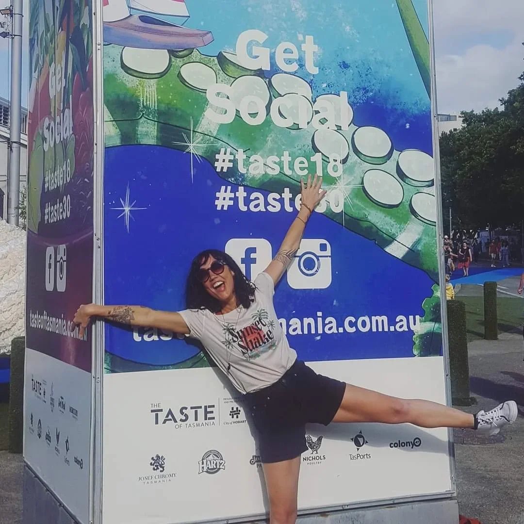 A woman posing playfully with outstretched arms and one leg lifted in front of a colorful outdoor advertisement for Taste of Tasmania, featuring social media hashtags and logos.