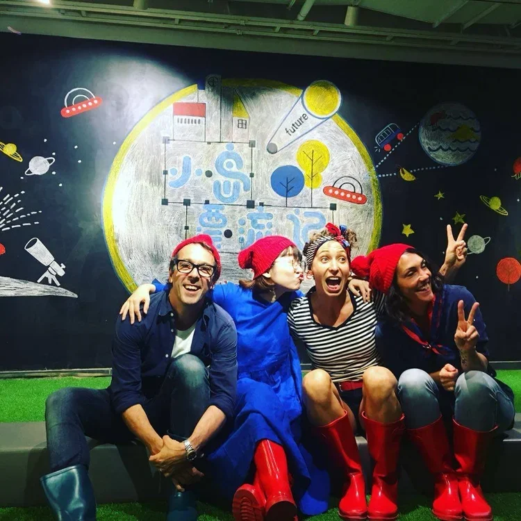 Four people sitting in front of a colorful space-themed mural, wearing red hats and rain boots, smiling, and making peace signs.