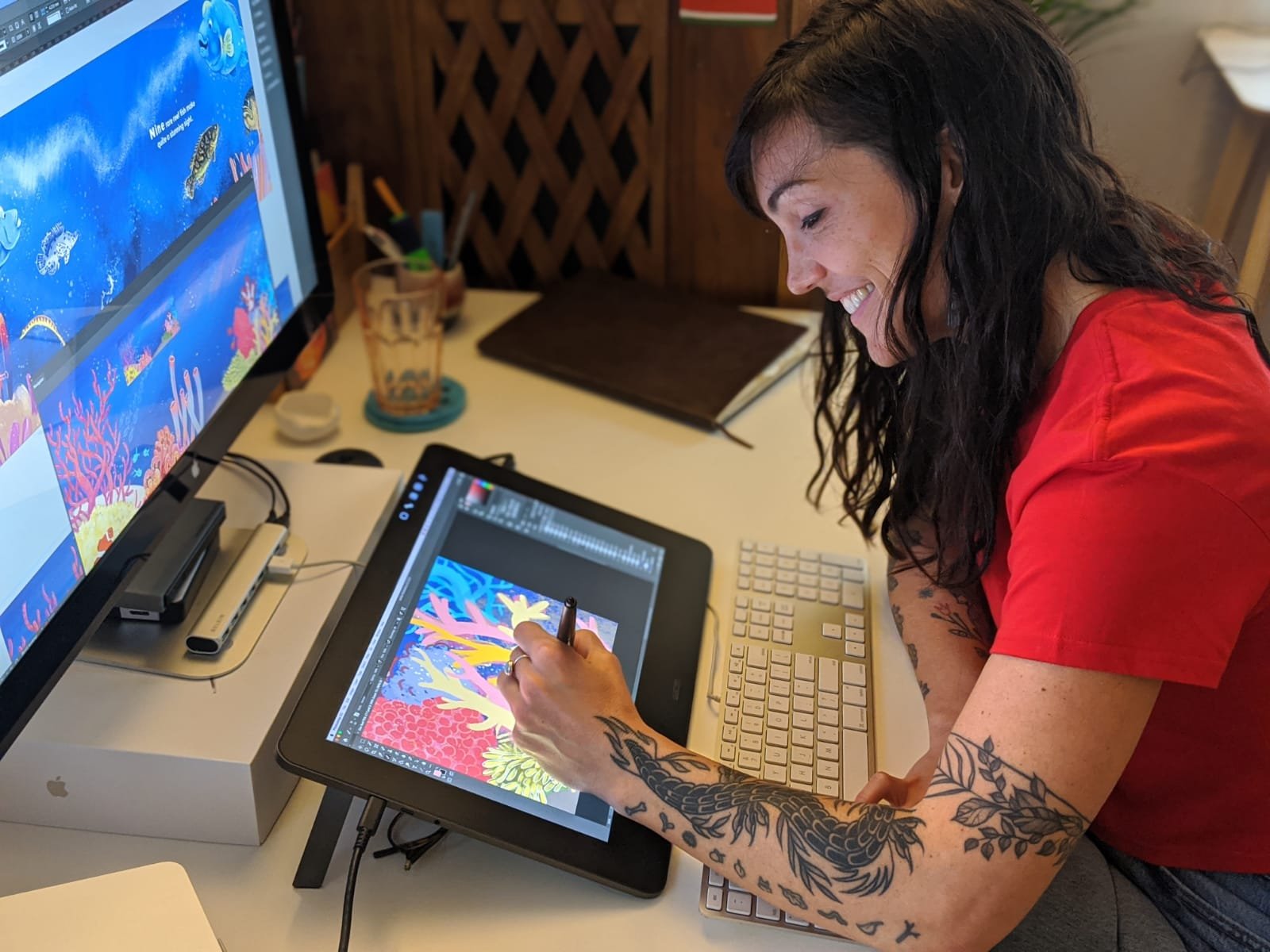 A woman with dark curly hair and tattoos on her arms is drawing a colorful underwater scene on a digital drawing tablet. She is smiling and working in a home office setting.
