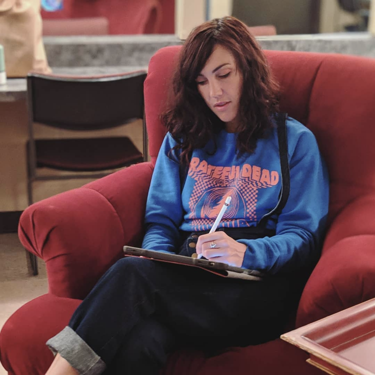 Woman with wavy brown hair sitting in a red armchair, writing in a notebook, wearing a blue graphic sweatshirt and black pants.