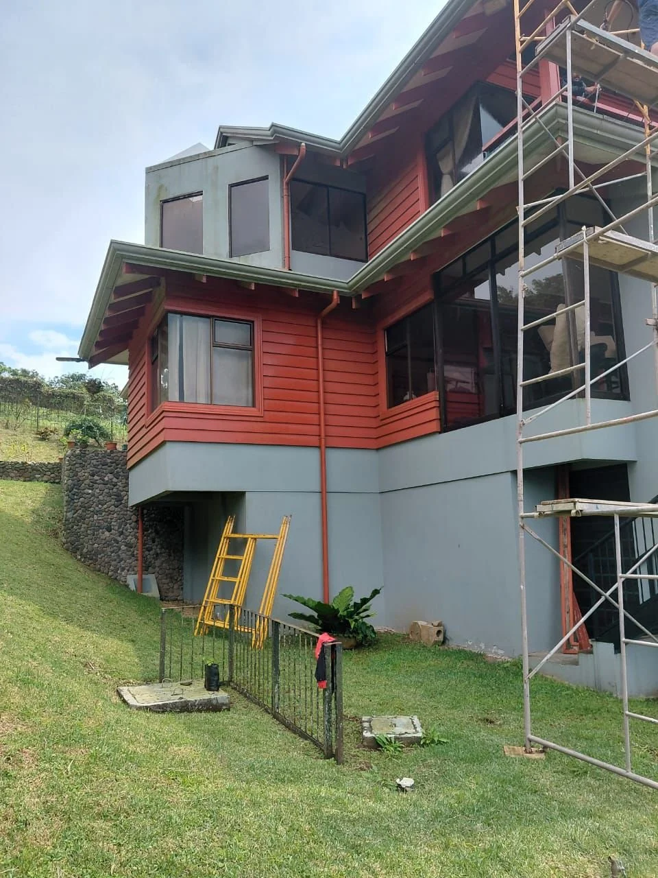 Construcción de una casa con estructura de madera y ventanas de cristal, con andamios y escaleras en el exterior, en un terreno con césped.