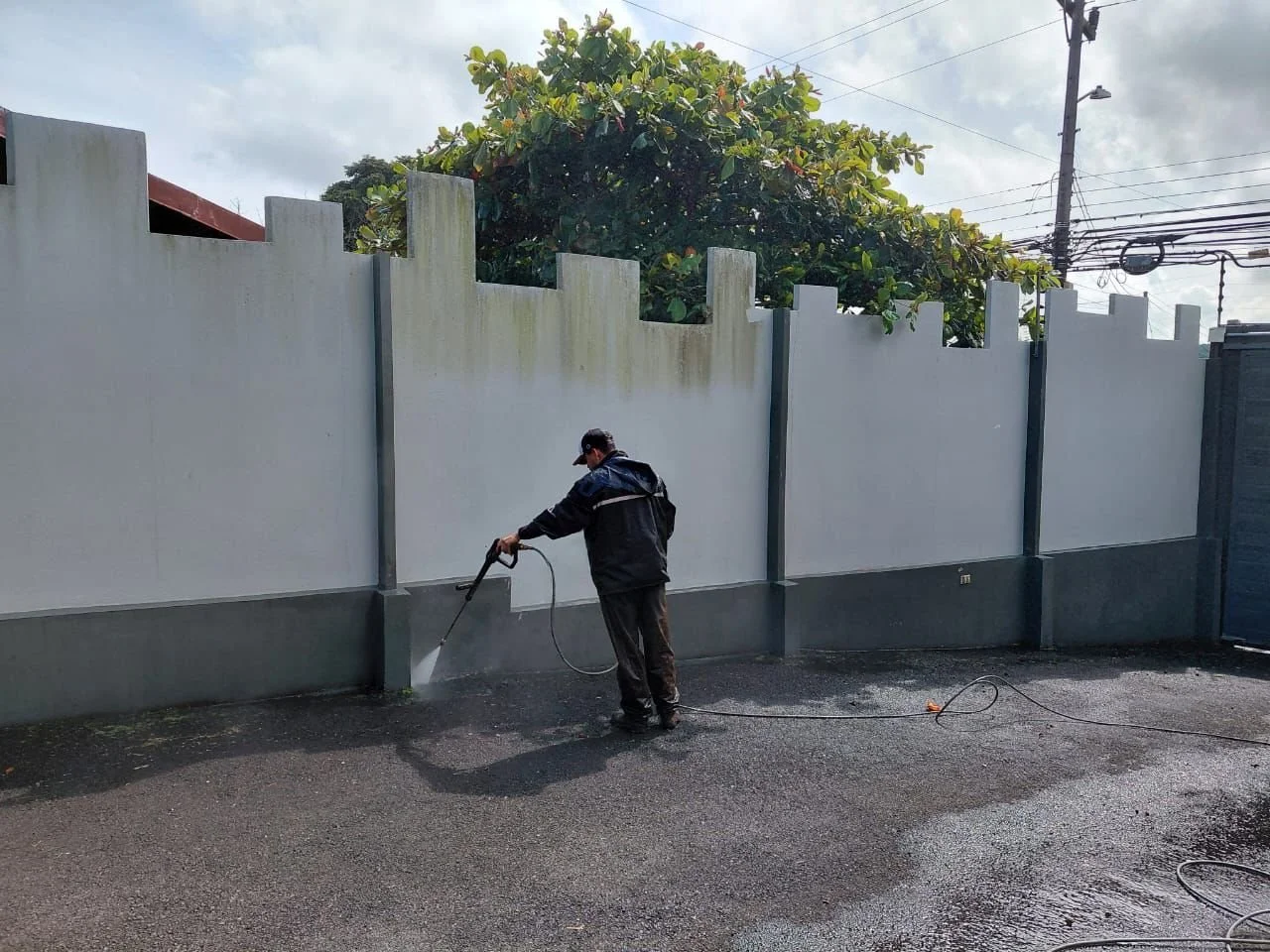 Hombre limpiando una pared con una pistola de agua en una calle asfaltada, con una cerca blanca y un árbol grande detrás, en un día nublado.