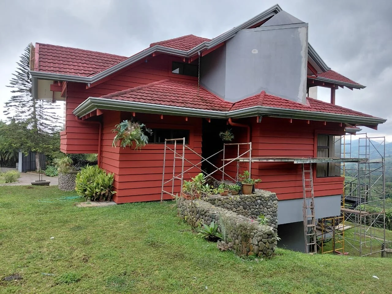 Casa en construcción con fachada de madera roja, techos de tejas rojas y andamios alrededor en un área de césped con plantas.