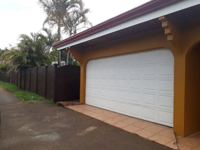 Vista frontal de un garaje con puerta blanca y marco de color amarillo, a la derecha de la casa, con una cerca de madera oscura a la izquierda y árboles de palma en el fondo.
