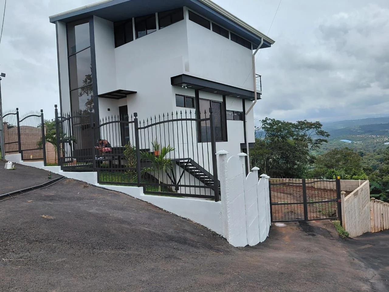 Casa moderna de dos pisos en una colina con vista panorámica, fachada blanca, ventanas grandes y barandales negros, rodeada de muro y portón de metal negro con paisaje verde.