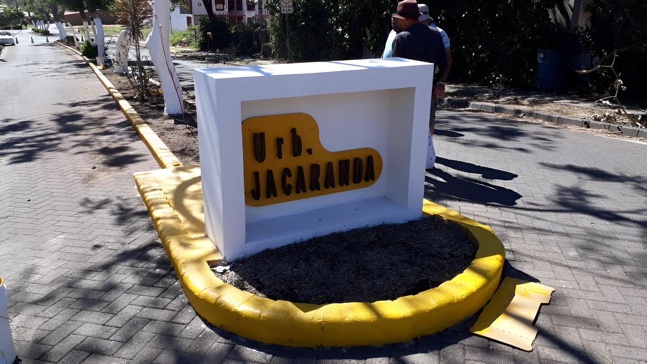 Se observa una señal de señalización en una calle, con letras en relieve que dicen 'urb. JACARANDA'. La señal está en una estructura blanca con borde amarillo, y está siendo colocada en una acera de piedra.