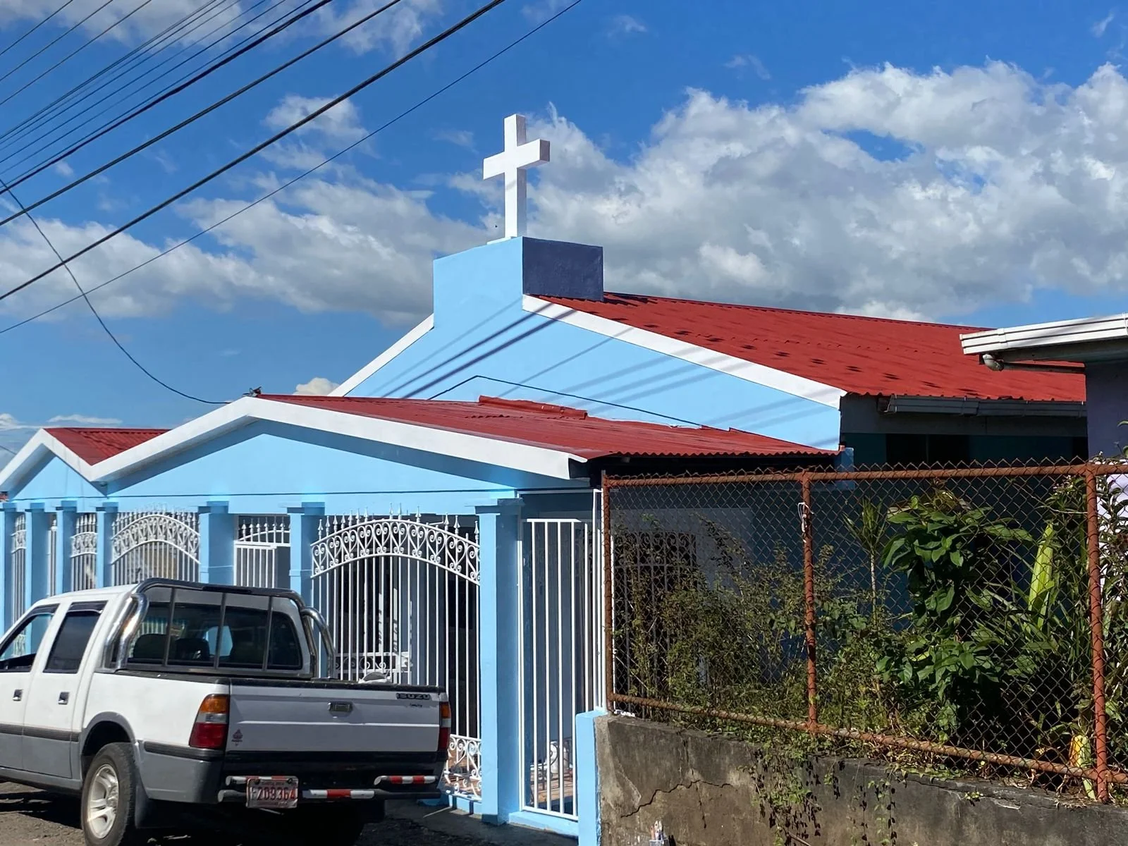 Una iglesia cristiana de color azul con una cruz blanca en el techo y un techo de metal rojo, al frente hay una cerca de metal blanca y un vehículo estacionado.